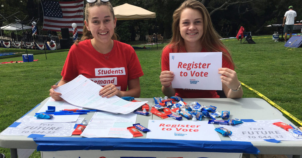 Two Students Demand Action volunteers smile while tabling at an outdoor event to register people to vote. On the table in front of the two volunteers are various forms, Students Demand Action signage, and candy