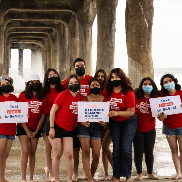 A group of ten people participating in the Summer Leadership Academy pose in Students Demand Action shirts on the beach in California.