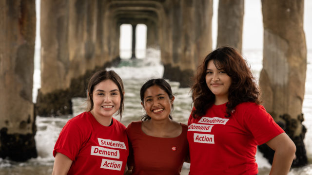 Three volunteers smile while wearing Students Demand Action shirts together on the beach