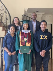 Ade smiles for a group photo with four of her family members at home. She wears a cap and gown and is holding her high school diploma.