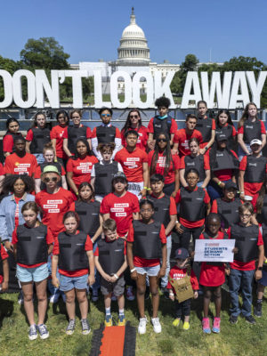 A group of students wearing bullet proof vests and Students Demand Action shirts stand in front of a sign that says Don't Look Away with the Capitol building in the background during an event in Washington, DC in June 2022