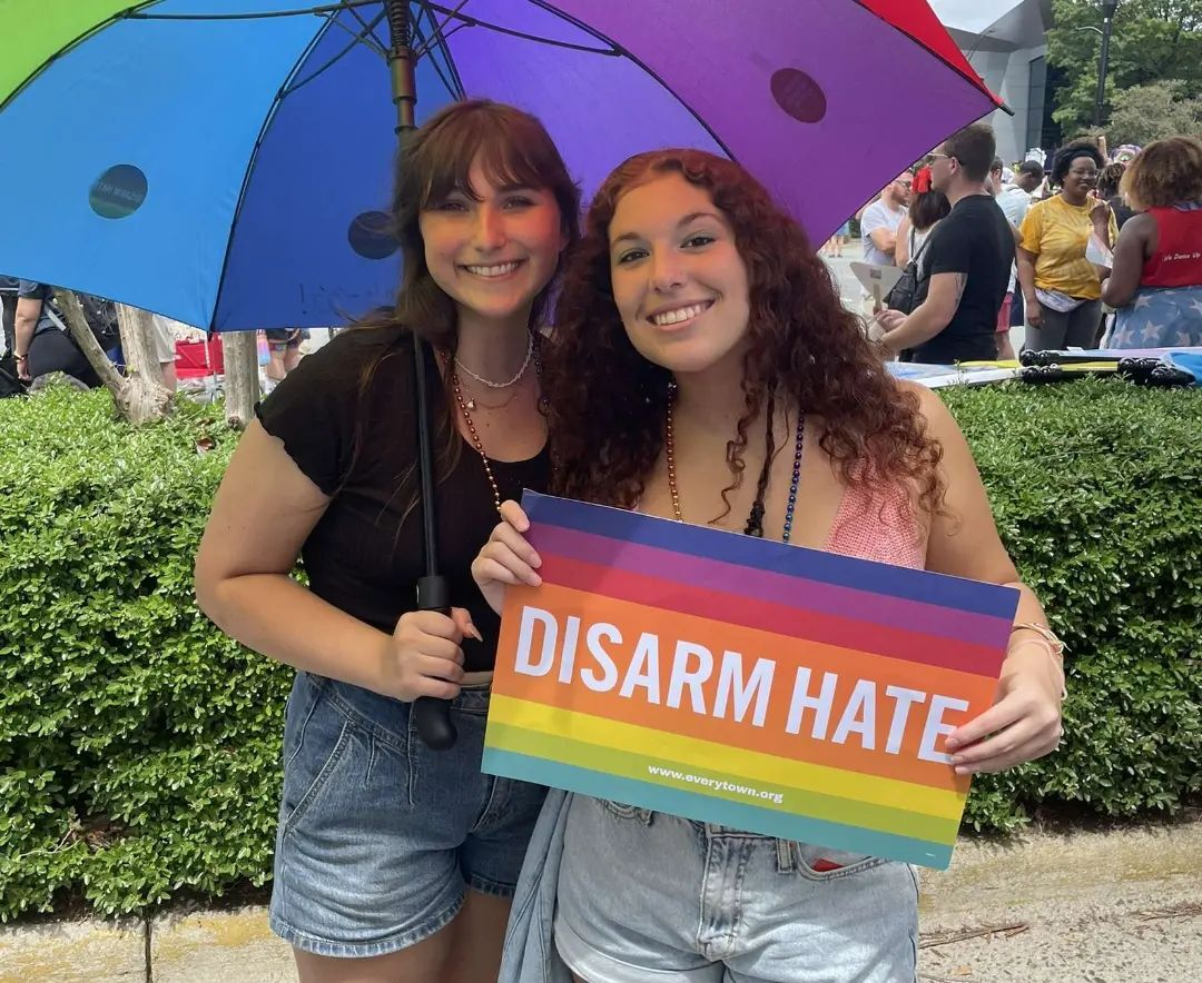 Two students posing for a photo, one of them holding a rainbow umbrella and the other holding a rainbow Disarm Hate sign