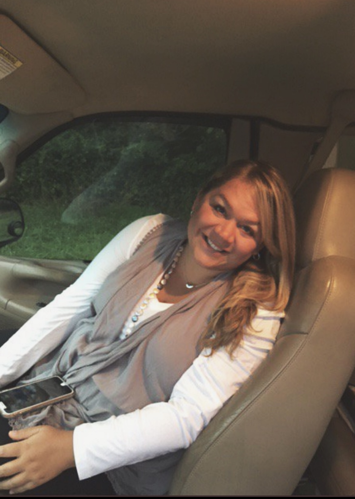 Pictured: Laura Wallen, a white woman with brownish-blond hair. She wears a white long-sleeve shirt, a flowy grey vest, and a long beaded necklace. She is smiling at the camera while leaning against the passenger seat of a vehicle; her iPhone sits on her lap.
