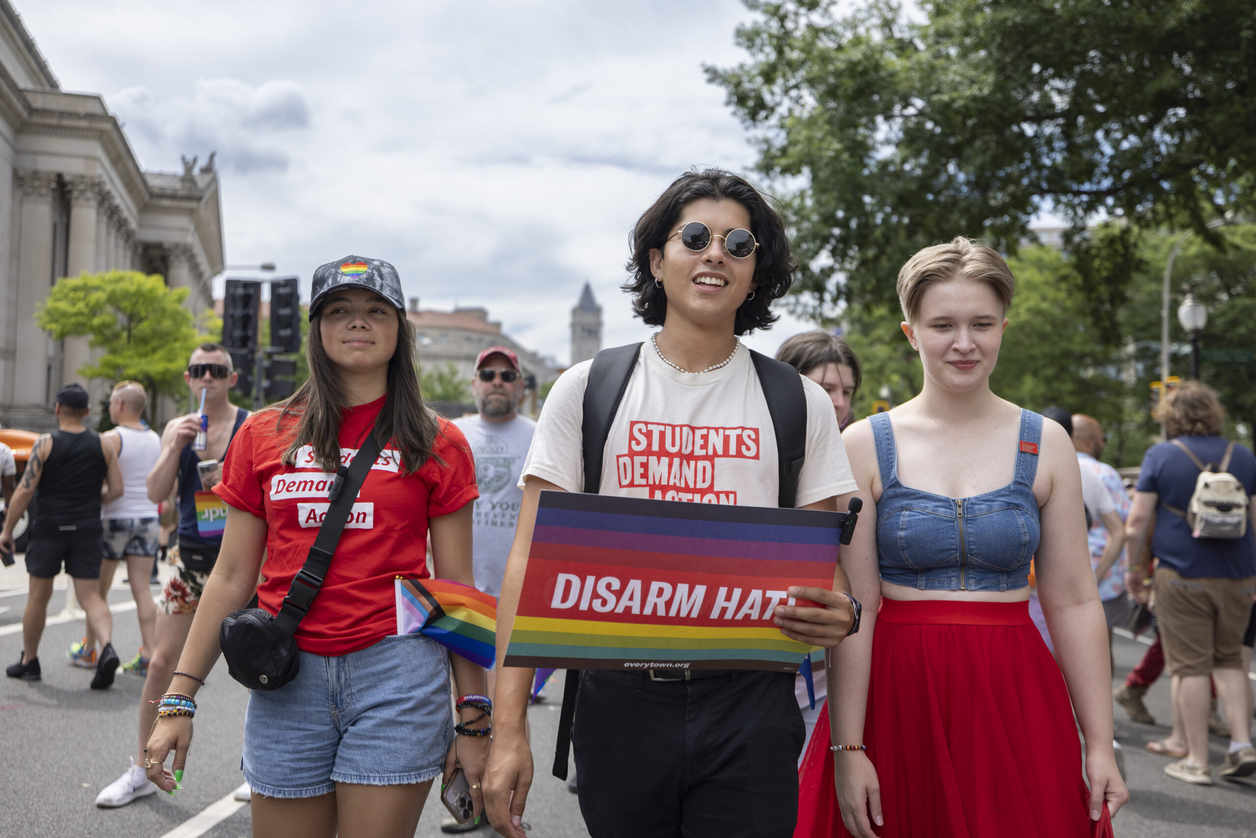 Andres Cubillos is in the center of the photo, marching in a DC Pride parade. He has curly dark hair cut to just above his shoulders and is wearing black round sunglasses. He wears an off-white t-shirt with Students Demand Action printed in all-caps in red/orange text. He holds a Disarm Hate sign (rainbow print background, with white text) in his left hand.