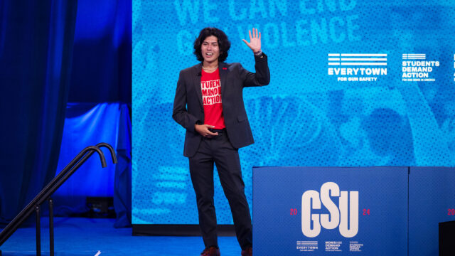 Andres Cubillos has curly dark hair cut just above his shoulders. He is standing against a blue background and wears a black suit with a red students demand action t-shirt. His left hand is raised in a wave.