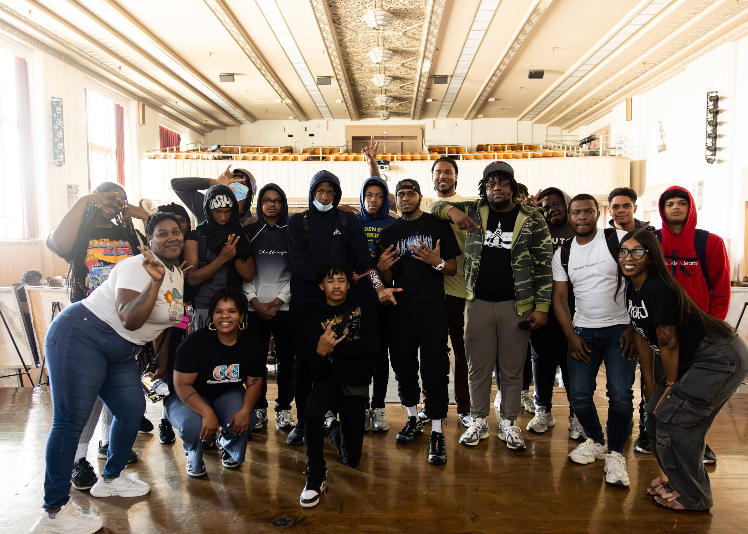 19 planning members of the conflict resolution tour gather on stage. Manny is pictured in the center wearing a black cap, black t-shirt, black pants, white socks, and sneakers.