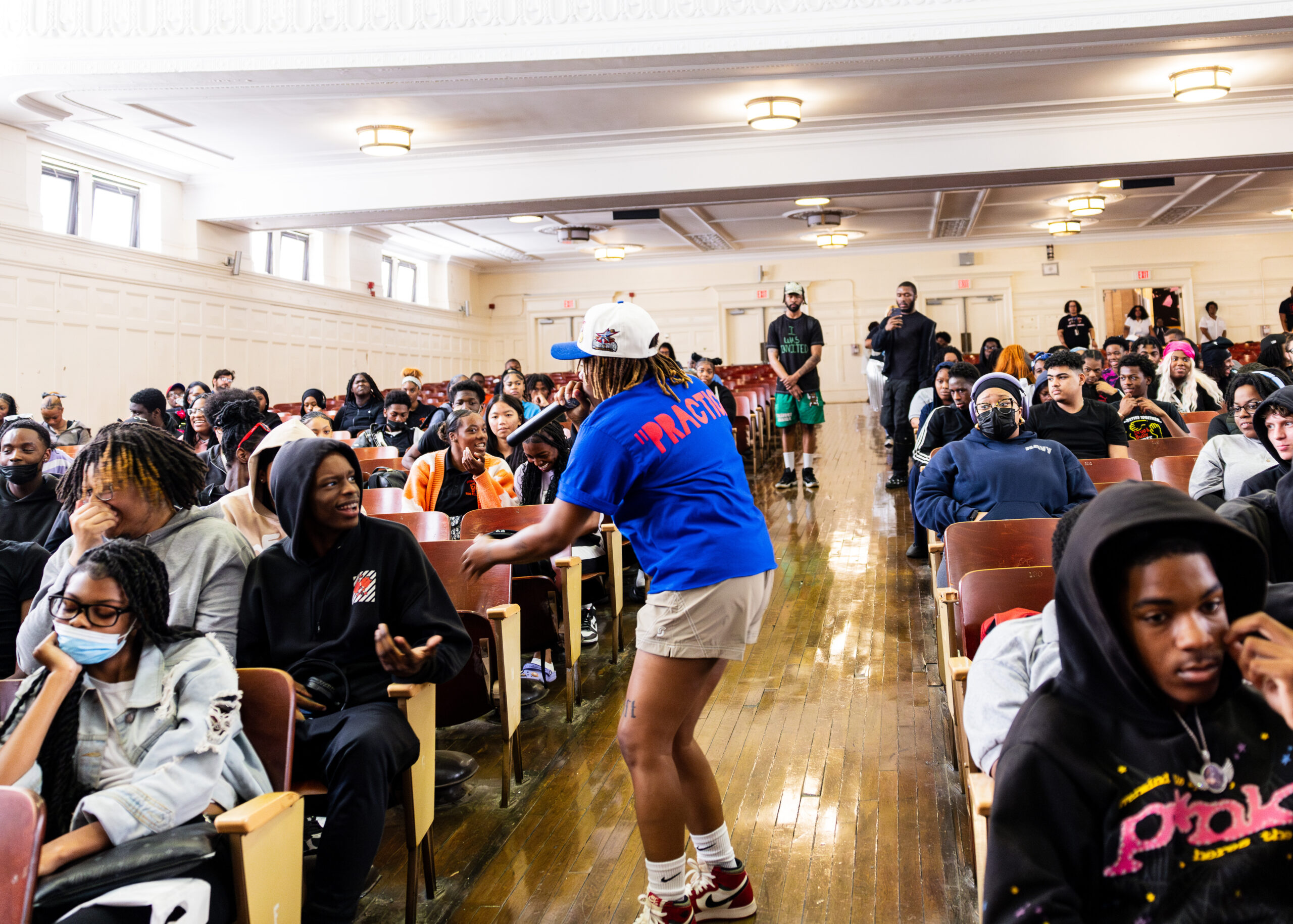 A Philadelphia artist performs at the Don't Get Tricked Out Ya spot tour. They are seen at a side profile and are wearing red and white Nike shoes, khaki pants, a cobalt blue shirt with neon orange lettering, and a white-and-blue baseball cap. They have brown dreadlocks that reach just below their shoulders. Students surrounding the artist are watching them perform.