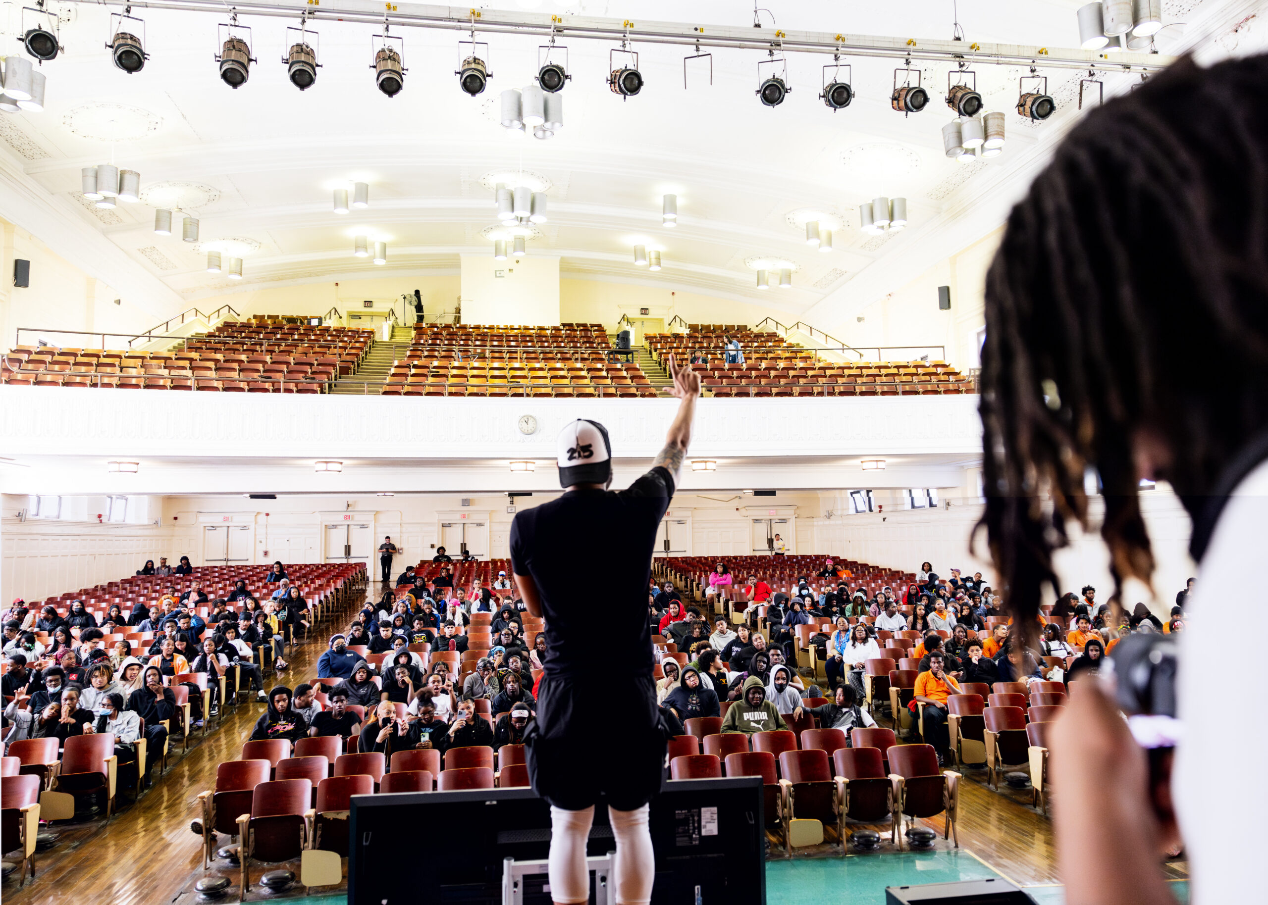 Manny raises his right arm as he talks to students attending the conflict resolution tour at Overbrook High School. The photo is taken from the back of the stage, showing Manny's back and the faces of many of the audience members. Manny wears black shorts, a black t-shirt, and a white-and-black paneled baseball cap that reads "215" in black lettering. In the right side of the frame, a person is pictured taking a photo of Manny speaking to the students.