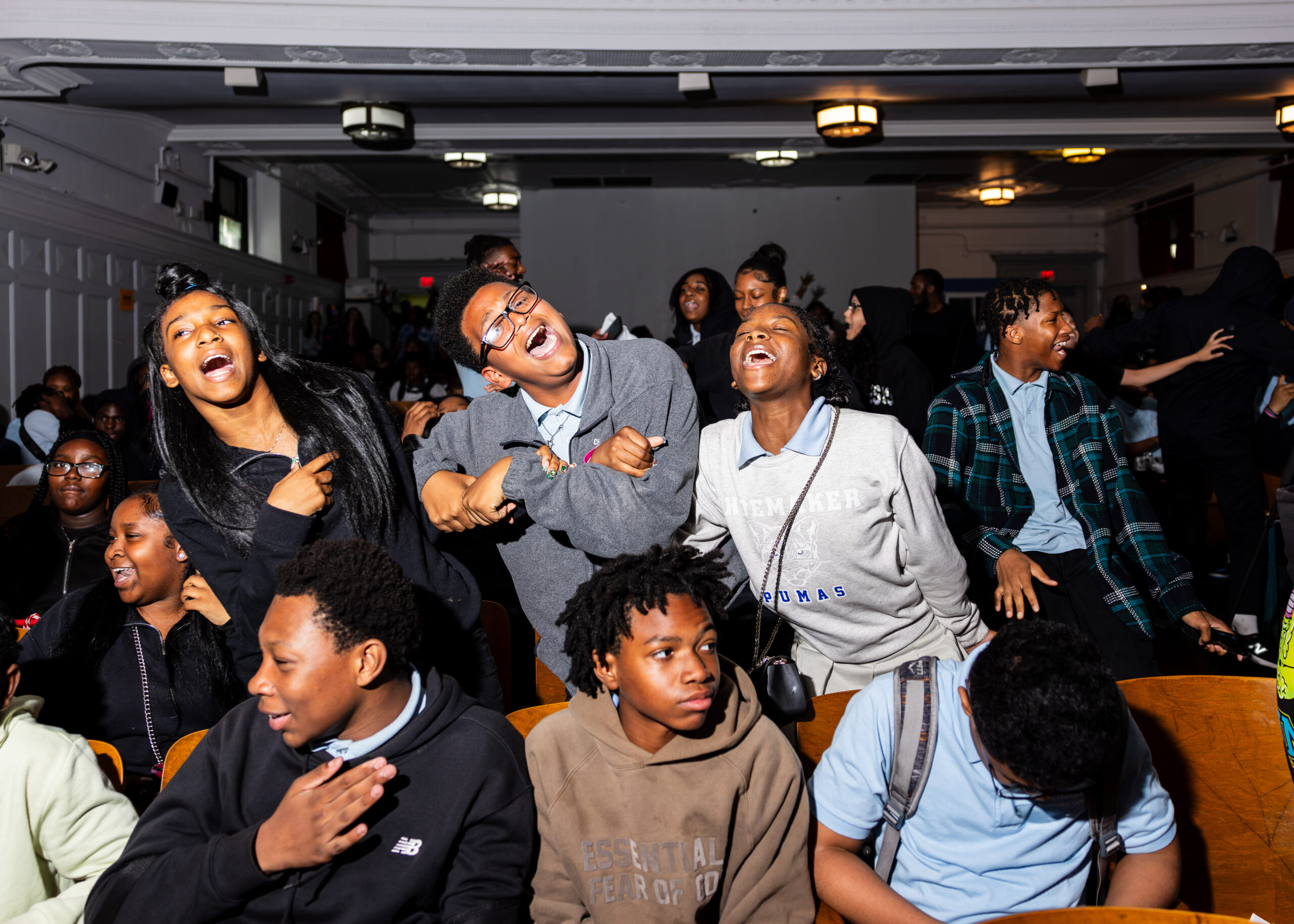 Three Black students are centered in the image, linking elbows and singing along to music. They are swaying and smiling with open mouths at the camera. Three other students are pictured in front of them, and many students in the background of the image are also dancing.