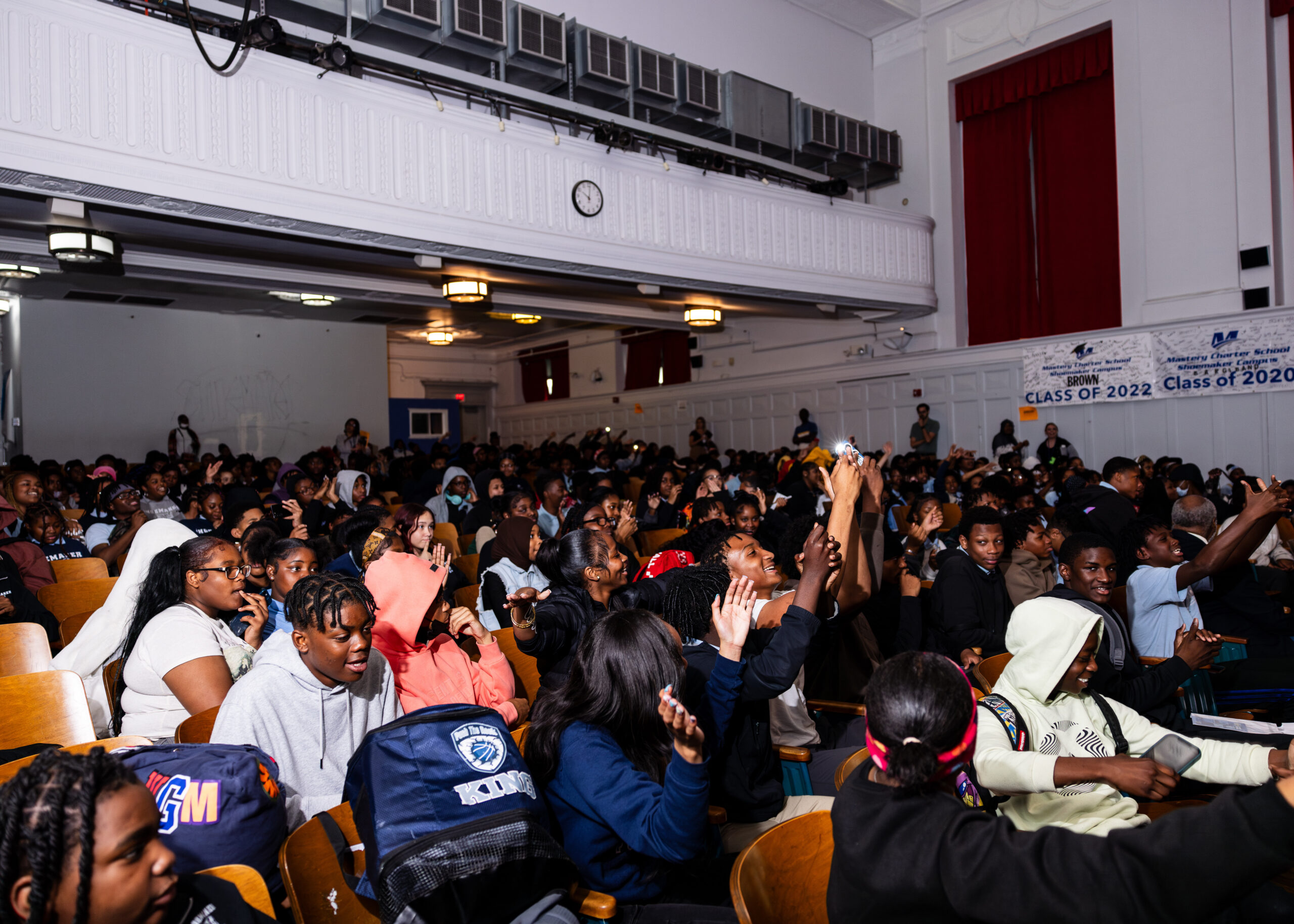 A photo taken at a slight profile angle of Black students attending the conflict resolution tour. The photo appears to have been taken with a camera flash. Many students are smiling and filming the stage.