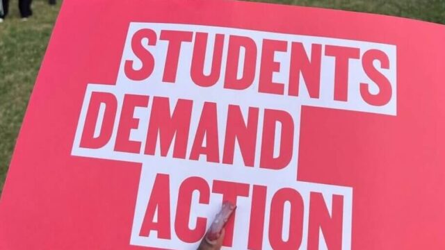 A red Students Demand Action sign is in focus in the foreground. In the background, students stand in a half-circle in a green space on campus.