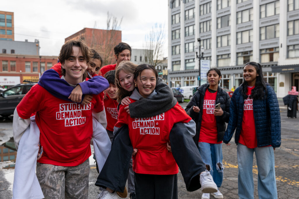 Students Demand Action volunteers pose for a group photo. Two students are being piggy-backed by two others. All are wearing red Students Demand Action t-shirts.