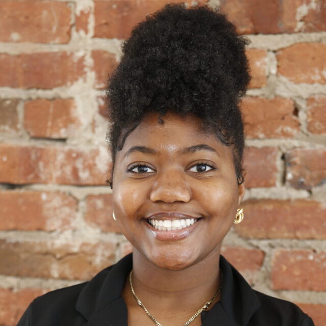 Iyanah Forbes, an HBCU student and the chartering president of Students Demand Action, poses for a professional photo. She stands in front of a brick wall and wears a black blazer, a gold necklace with a pendant, and small dangly gold earrings.