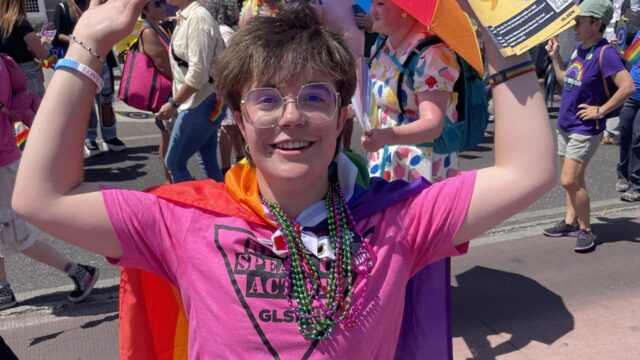 Bo, an SDA leader, wears a pink t-shirt, a red cape, and is holding pride flags at a Pride event.