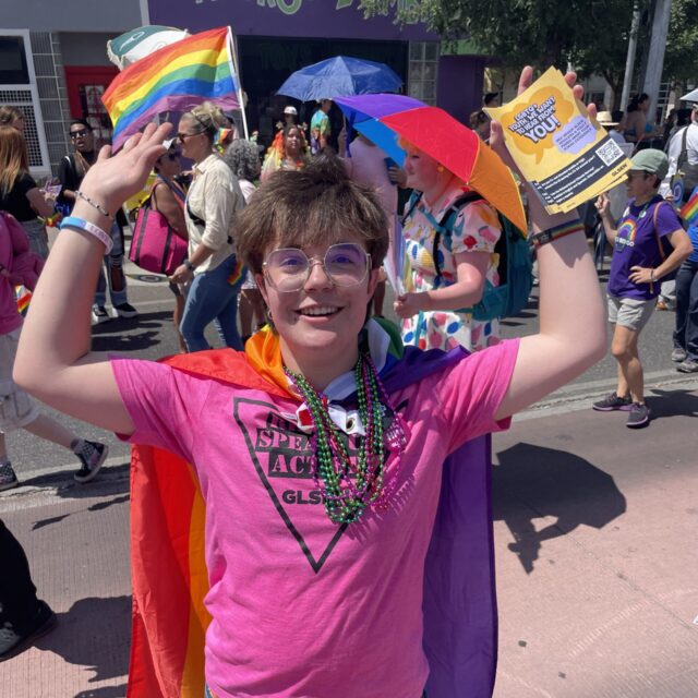 Bo, an SDA leader, wears a pink t-shirt, a red cape, and is holding pride flags at a Pride event.