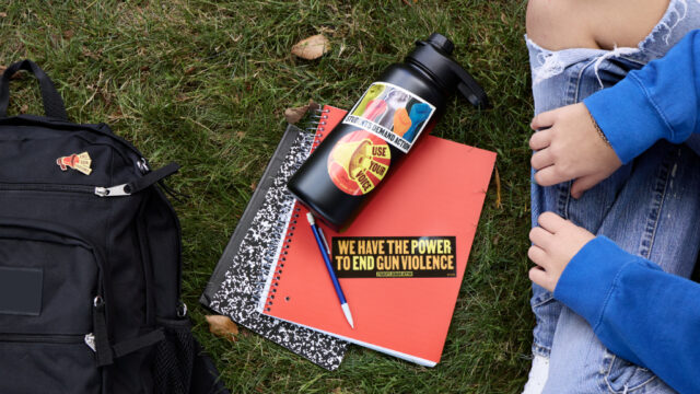 A person sits cross-legged on grass. A black backpack with a Students Demand Action megaphone pin, a black water bottle with a Students Demand Action megaphone sticker, and a red notebook with a Students Demand Action