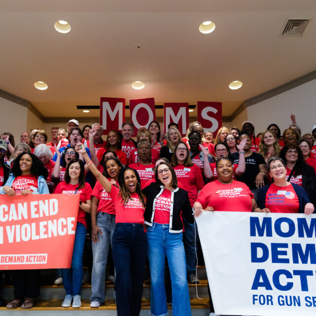 A large group of people, all wearing red Moms Demand Action or Students Demand Action t-shirts, are smiling, clapping, and cheering in a group photo. In the front left, a group of people hold a banner that says