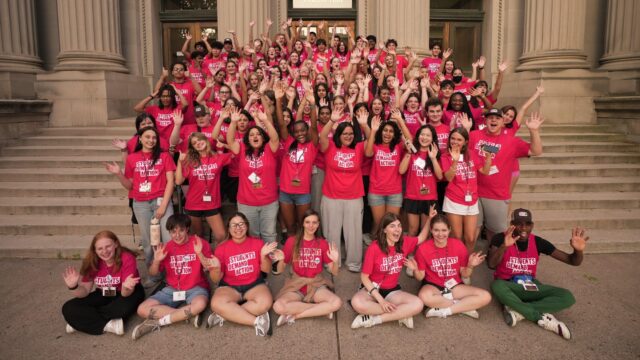 A group of 100 Students Demand Action volunteers, all wearing red t-shirts with the Students Demand Action logo, gather for a photo on the Vincent Hall steps at the University of Minnesota during the Summer Organizing Institute. Students are raising their arms in the air and waving at the camera.