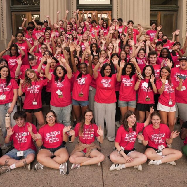 A group of 100 Students Demand Action volunteers, all wearing red t-shirts with the Students Demand Action logo, gather for a photo on the Vincent Hall steps at the University of Minnesota during the Summer Organizing Institute. Students are raising their arms in the air and waving at the camera.