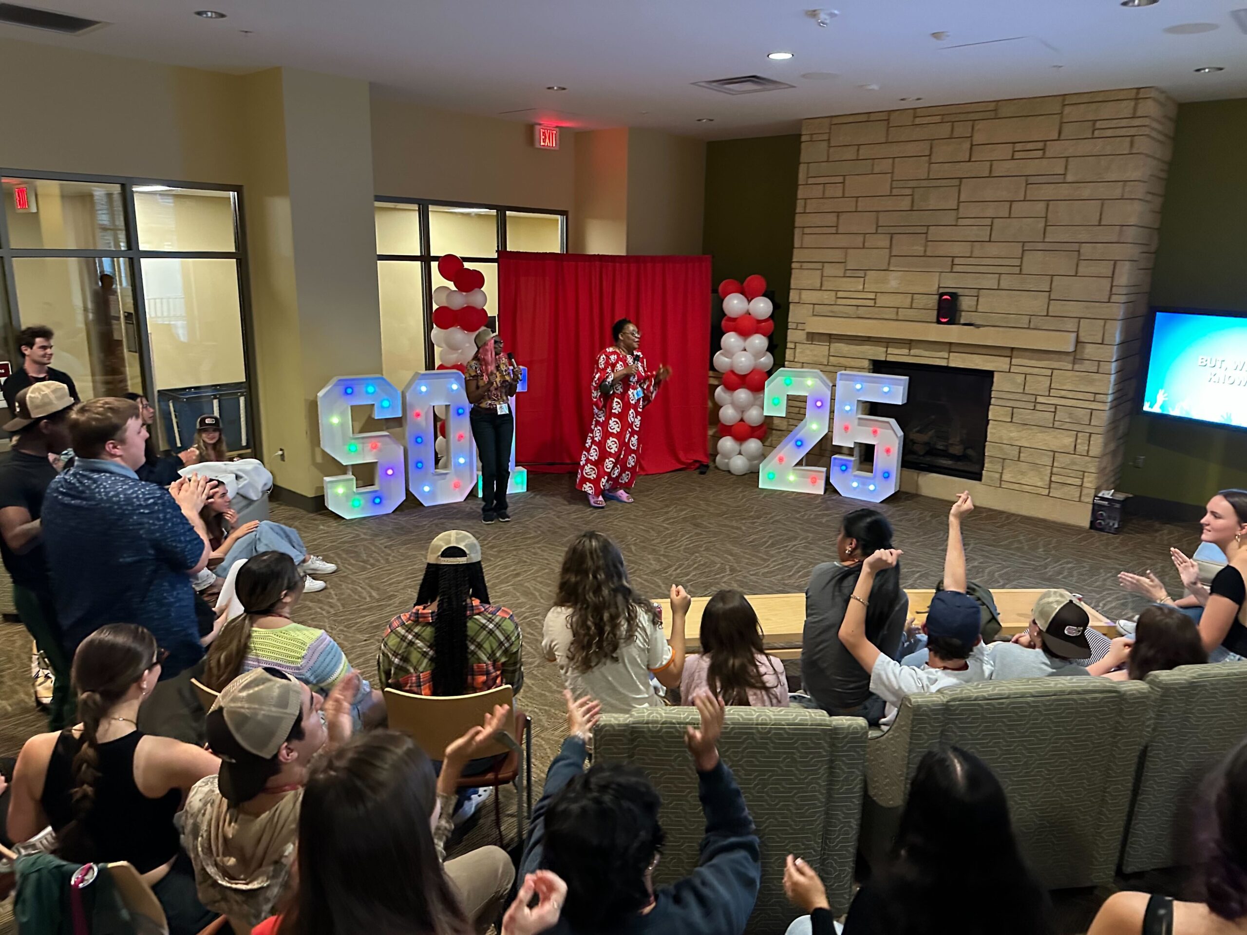 A group of students and staff gather in a University of Minnesota dorm common area to sing karaoke during the Students Demand Action Summer Organizing Institute. Two staff members are singing, while a group of approximately 20 students sit in a semi-circle and cheer them on.