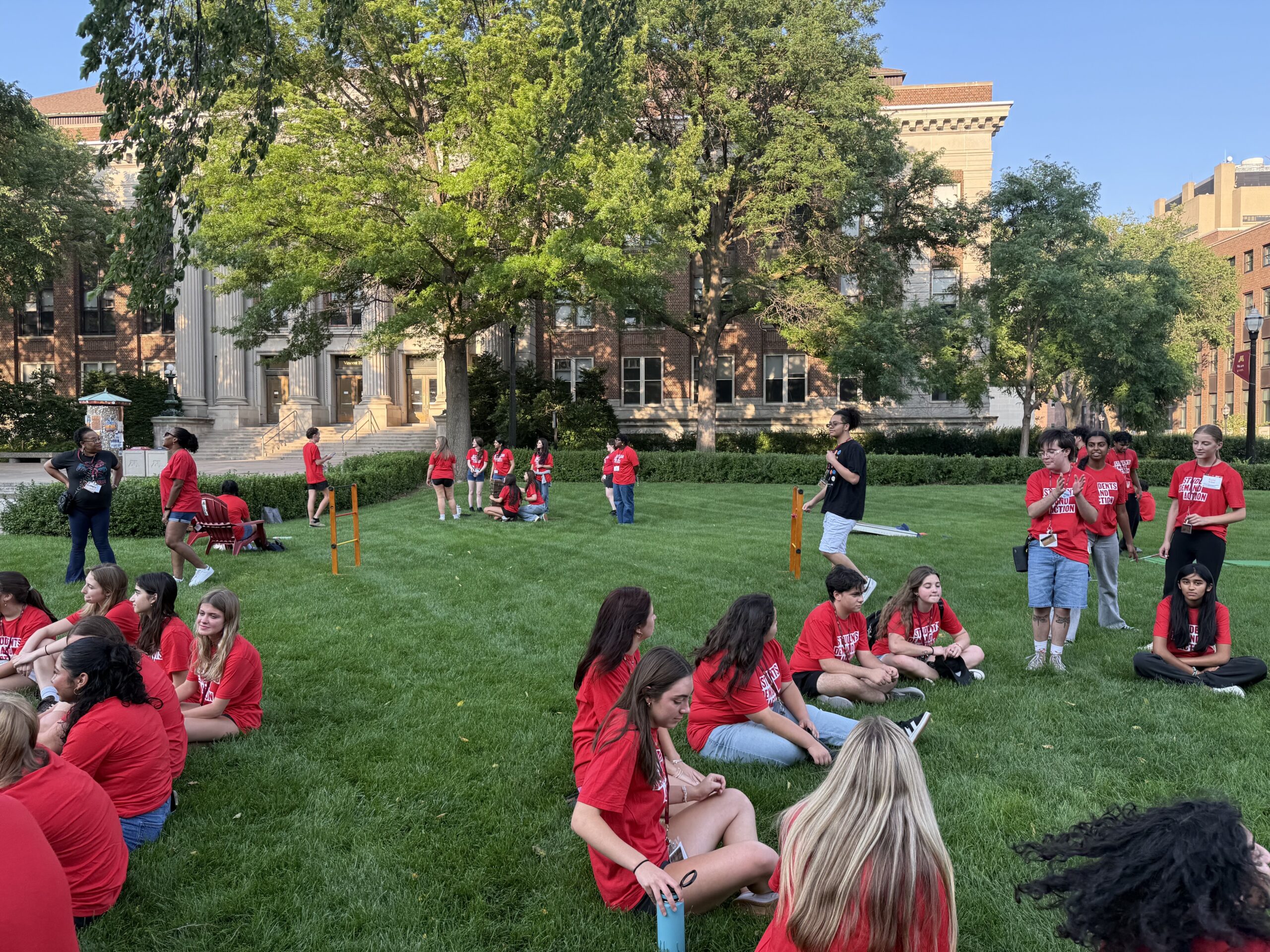 Students Demand Action Summer Organizing Institute attendees, all wearing red Students Demand Action t-shirts, sit or stand in small groups on the lawn to play games at the University of Minnesota.