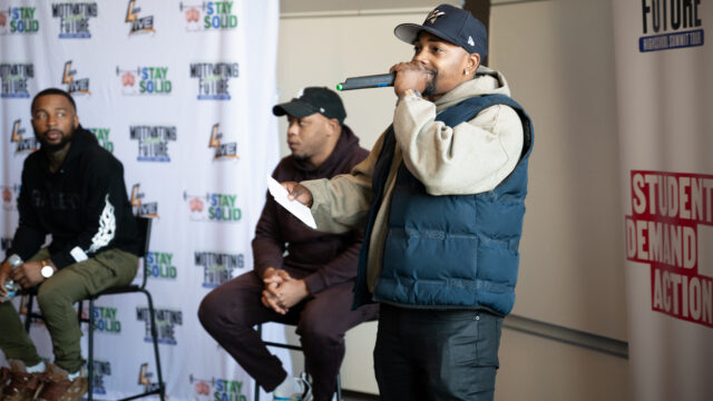Neal McDonald wears a black hat, tan hoodie, blue puff vest, and dark pants. He is speaking into a microphone to a crowd of students off camera. Behind him are two panelists on the Motivating Our Future tour.