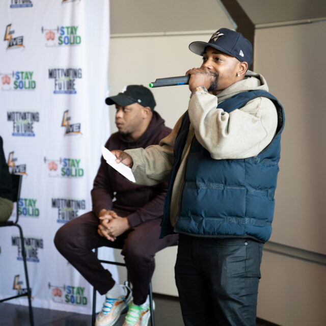 Neal McDonald wears a black hat, tan hoodie, blue puff vest, and dark pants. He is speaking into a microphone to a crowd of students off camera. Behind him are two panelists on the Motivating Our Future tour.