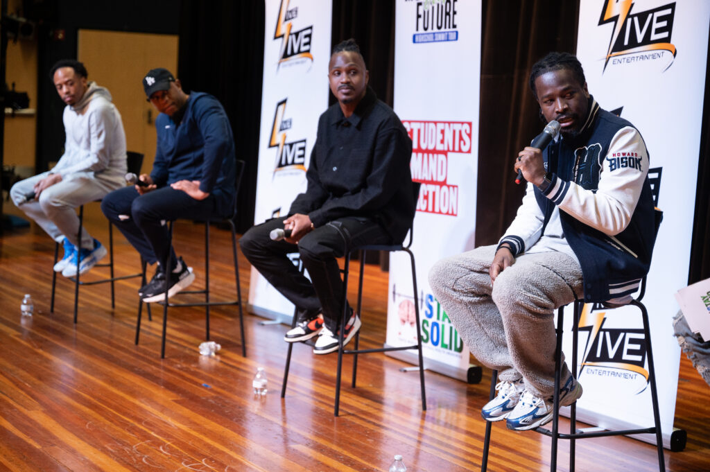 Panelists at the Motivating Our Future Summit Tour look to their left at a fourth panelist, who is holding a microphone and looking off camera as he speaks during the Motivating Our Future Summit Tour.