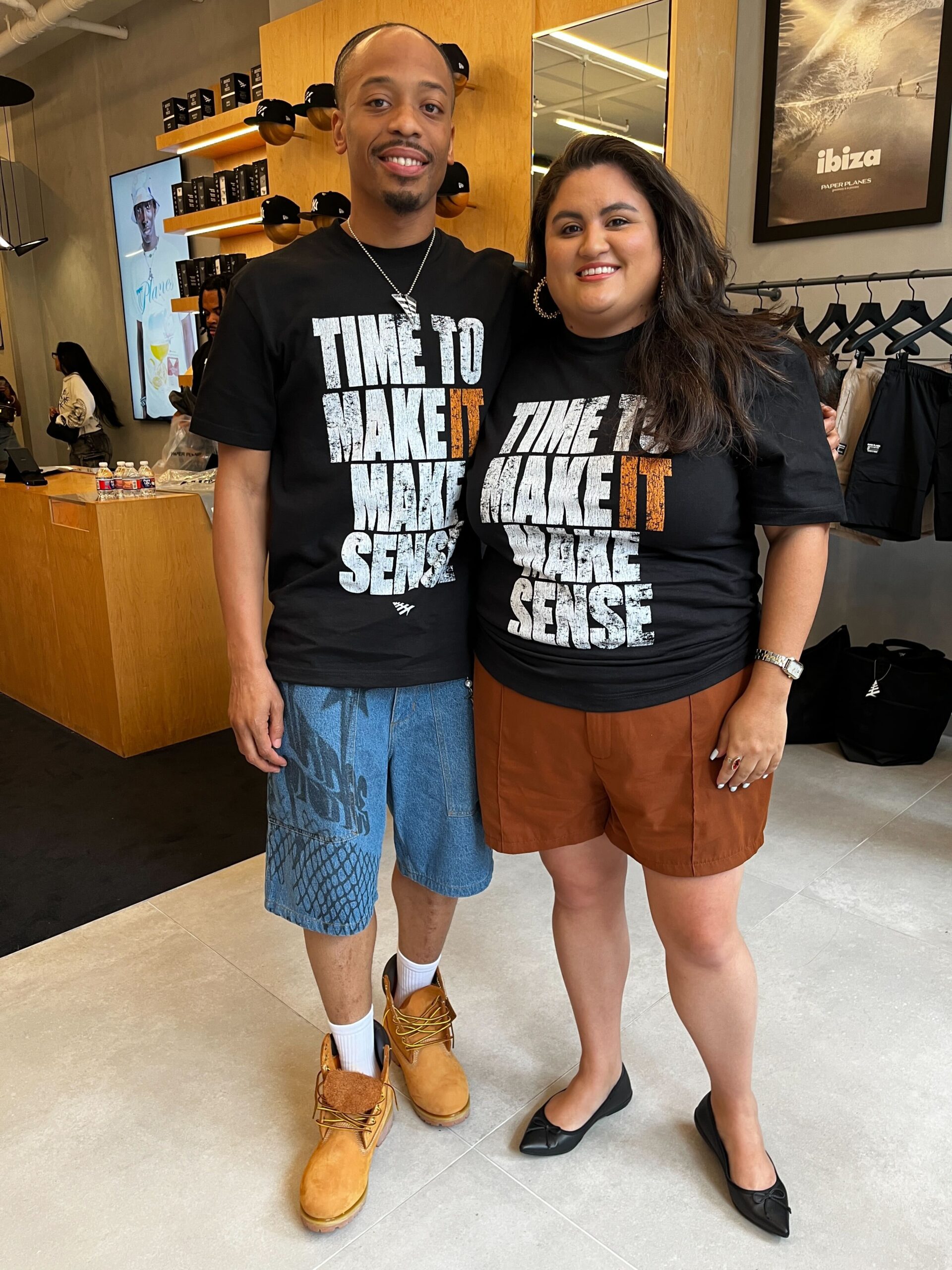 Justin Murray and Andrea Azalia pose for a photo at the Paper Planes pop-up event in honor of Neal McDonald. Both are wearing the black "Time to Make It Make Sense" t-shirt.