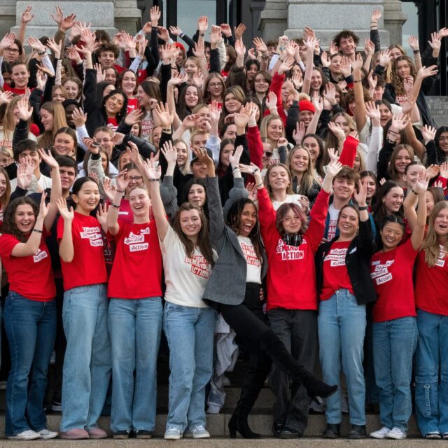 Angela Ferrell-Zabala, executive director of Moms Demand Action and Students Demand Action, stands on the steps of the Colorado State Capitol surrounded by over 100 Students Demand Action volunteers. Students are celebrating with their arms up in the air; many are wearing jeans and cream or red Students Demand Action t-shirts.