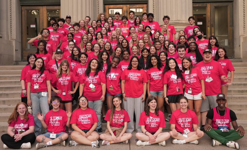 A group of 100 Students Demand Action volunteers, all wearing red t-shirts with the Students Demand Action logo, gather for a photo on the Vincent Hall steps at the University of Minnesota during the Summer Organizing Institute.