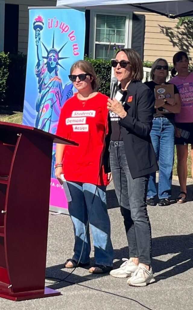 A Students Demand Action volunteer wears a red shirt and sunglasses; she stands behind a podium and is being introduced to an off-camera crowd by a Moms Demand Action volunteer beside her.