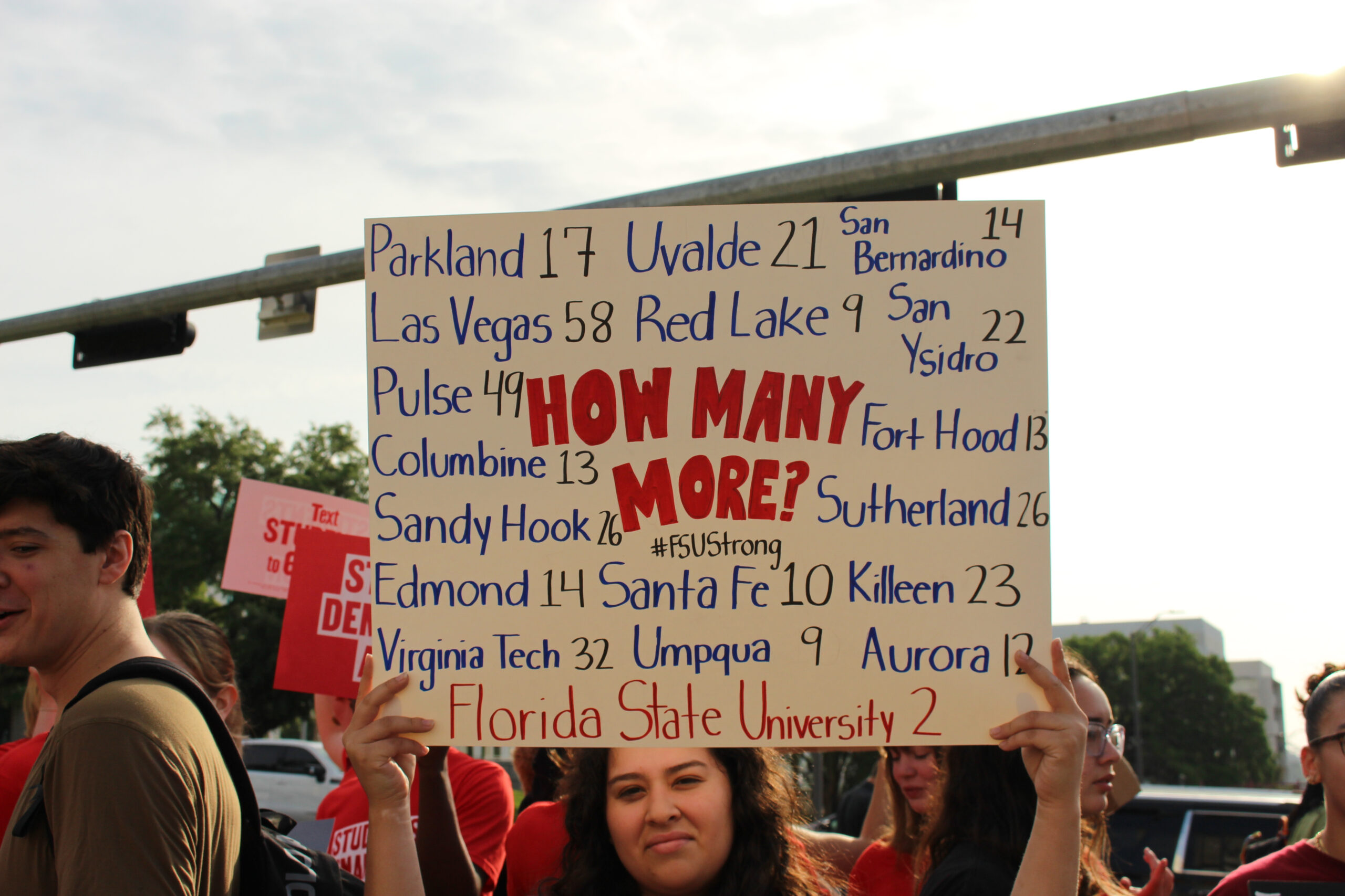 A protestor holds a sign that reads "How Many More?" in block red capital letters with the #FSUStrong written underneath the question. The sign also includes a list of names of places where mass shootings with a high number of casualties have taken place in the US: Parkland 17; Uvalde 21; San Bernardino 14; Las Vegas 58; Red Lake 9; San Ysidro 22; Pulse 49; Fort Hood 13; Columbine 13; Sutherland 26; Sandy Hook 26; Edmond 14; Santa Fe 10; Killeen 23; Virginia Tech 32; Umpqua 9; Aurora 12; Florida State University 2.