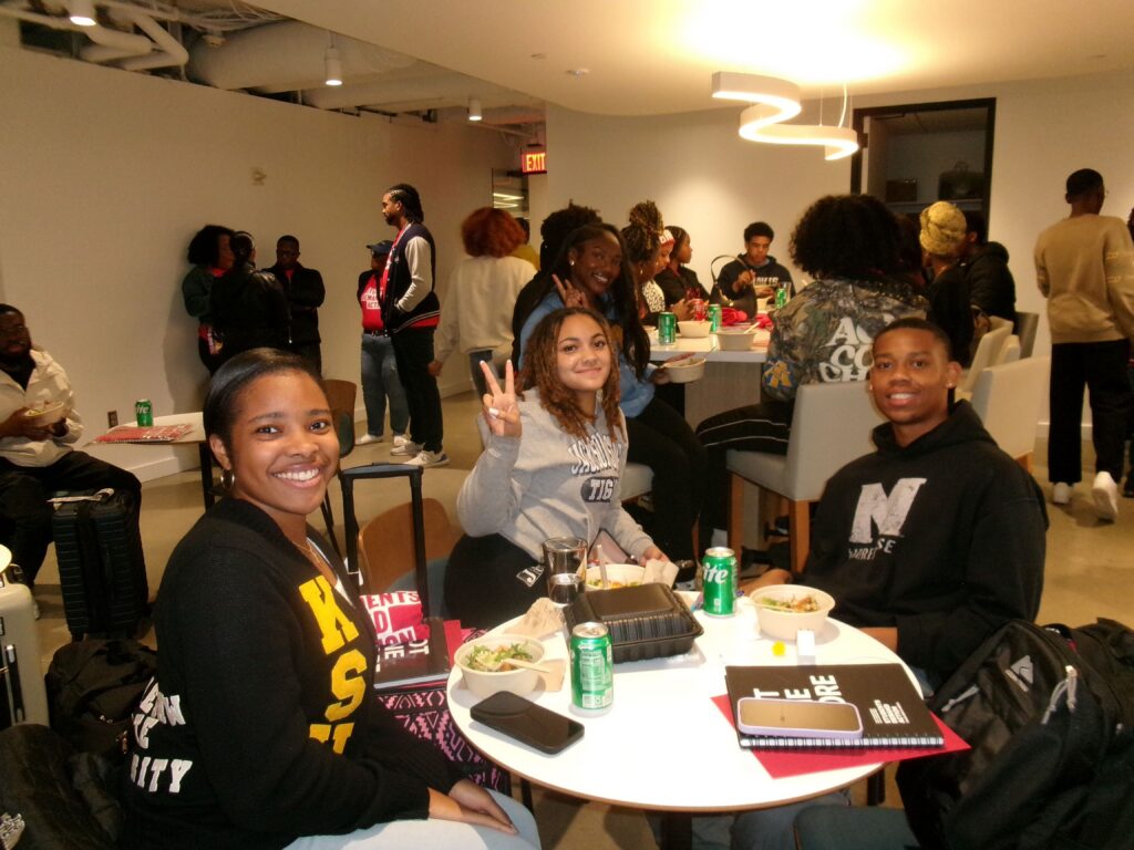 Three students pose for a photo at the Young Changemakers Summit. Approximately 20 other people are visible in the background.