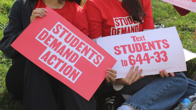 Two students are sitting in the grass holding signs. One reads 